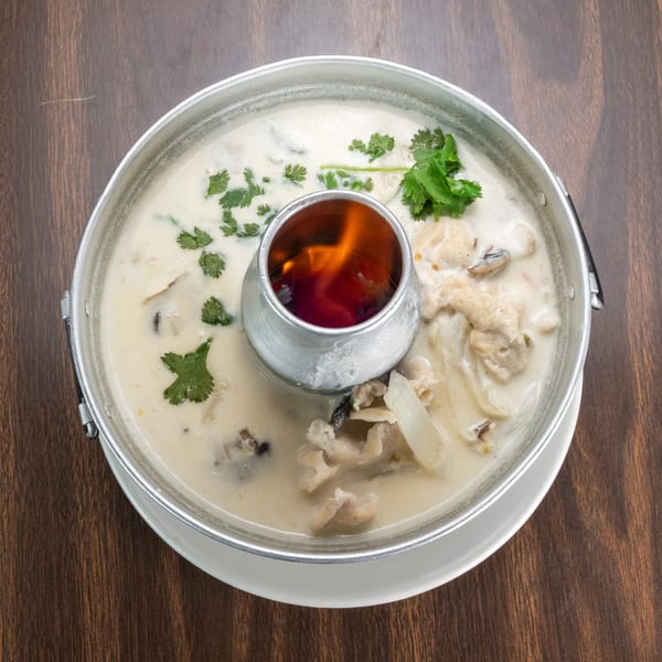 Creamy Tom Kha Gai soup with chicken, mushrooms, and cilantro in a hot pot with a flame.